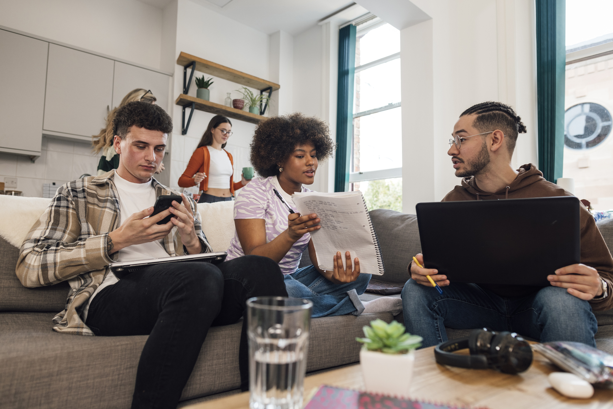 A low angle view of a group of teenage students who are smiling and working together in the common room. They are having social time and also catching up with some work together. They all have various wireless technology including laptops and headphones.